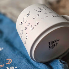 Load image into Gallery viewer, White mug with black text on a blue fabric background