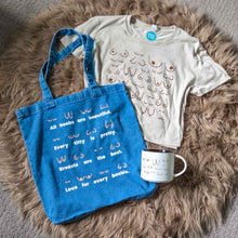Load image into Gallery viewer, Blue tote bag, white t-shirt with graphic design, and white mug on a fluffy brown rug.