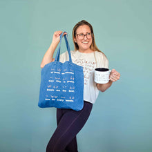 Load image into Gallery viewer, Person holding a blue tote bag and a white mug with text on a teal background