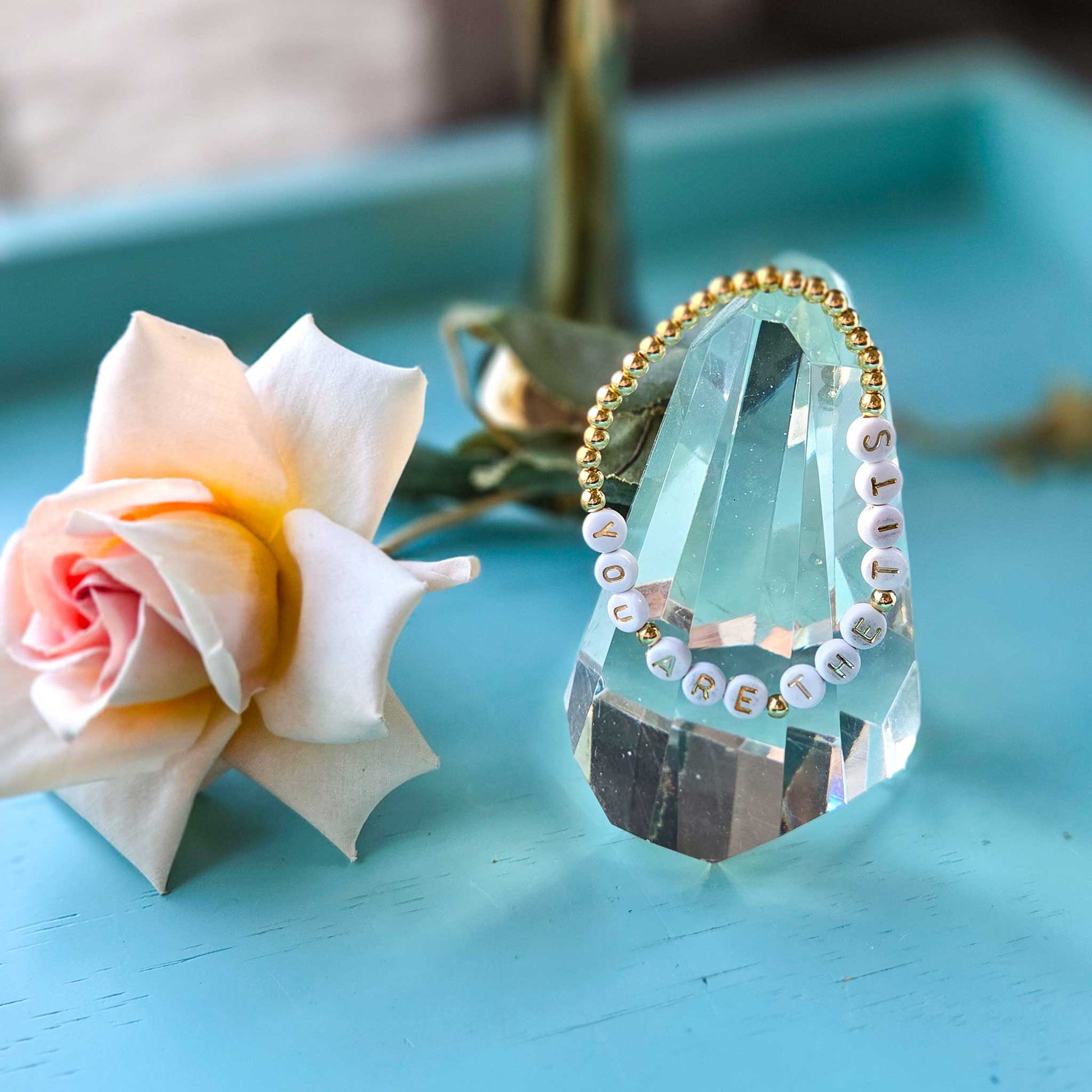 Crystal decoration with bracelet with text next to a pink flower on a blue surface