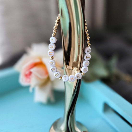 Gold hoop bracelet with white beads spelling 'YOU ARE THE TITS' on a stand with a blurred floral background.