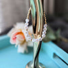 Gold hoop bracelet with white beads spelling 'YOU ARE THE TITS' on a stand with a blurred floral background.