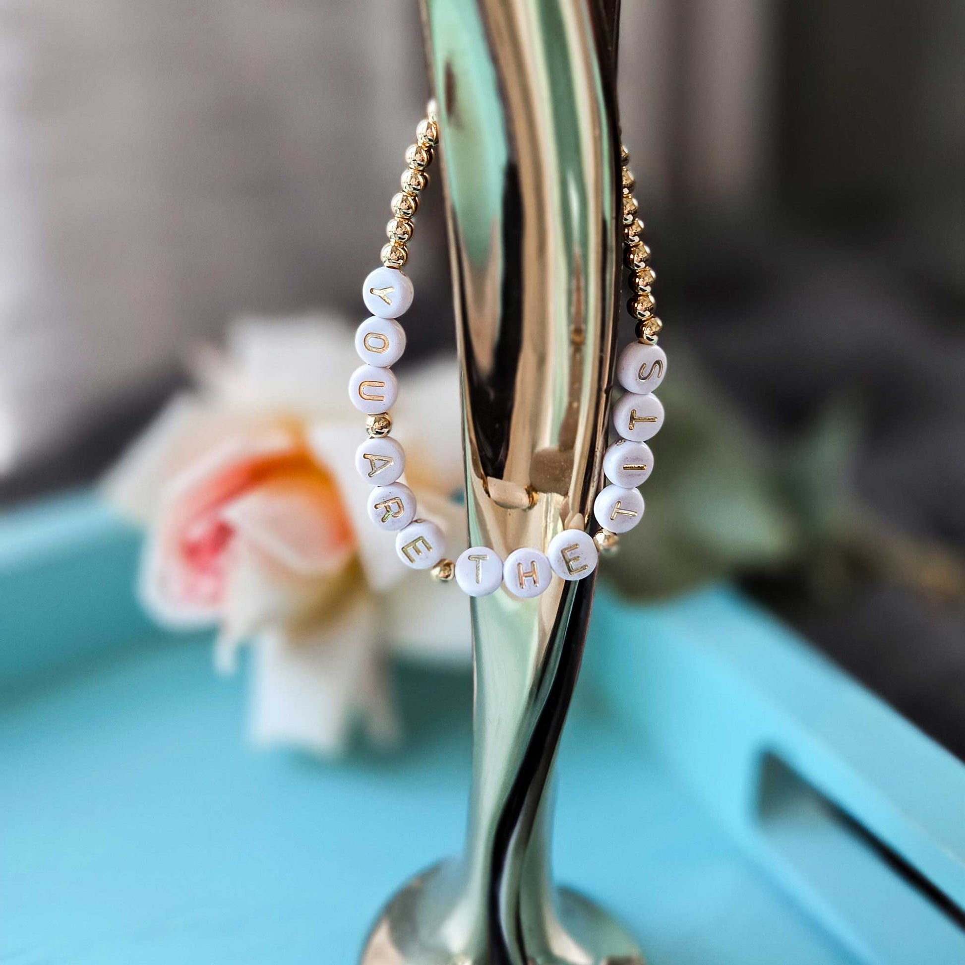 Gold hoop bracelet with white beads spelling 'YOU ARE THE TITS' on a stand with a blurred floral background.