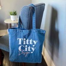 Load image into Gallery viewer, Blue tote bag with 'Titty City Design' text hanging on a chair indoors.
