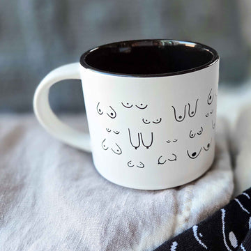 White mug with black interior and decorative boob print pattern on a soft fabric background