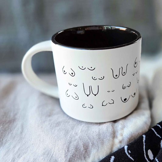 White mug with black interior and decorative boob print pattern on a soft fabric background