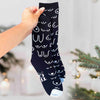 A hand holds up a black crew sock with a white novelty pattern of stylized boobs and cleavage motifs, set against a softly blurred holiday background with a Christmas tree.