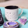 White mug with black designs on a blue surface with jewelry and crystals.
