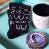 Black socks with white patterns, a white mug, and a purple coaster with bracelets on a teal surface.