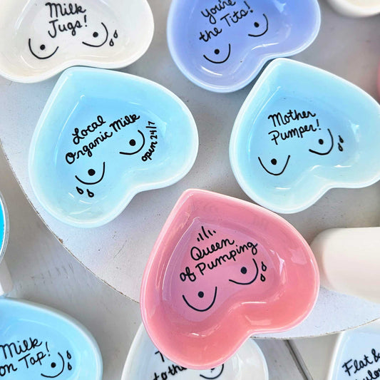 A set of heart-shaped ceramic dishware in pastel colors, including pink, light blue, lavender, and white, each decorated with a simple smiley face and humorous pregnancy-themed labels like 'Queen of Pumping' and 'Local Organic Milk'.