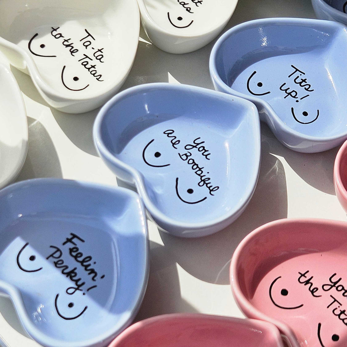 A collection of small heart-shaped ceramic dishes in pastel blue, pink, and white, each decorated with a hand-drawn smiley face and a playful handwritten phrase inside.