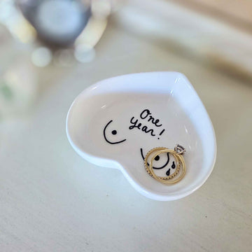Small white dish with a gold ring inside, featuring the text 'One year!' on a light wooden surface.