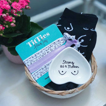 Titty City Design gift bundle with a white Strong as a Mother heart-shaped jewelry dish, black boob socks, and patterned hair ties displayed in a basket.
