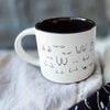 White mug with black interior and decorative text pattern on a soft fabric background