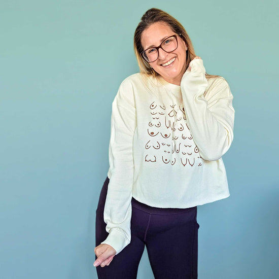 Woman wearing a cream self-love crop sweatshirt with design and dark pants against a light blue background