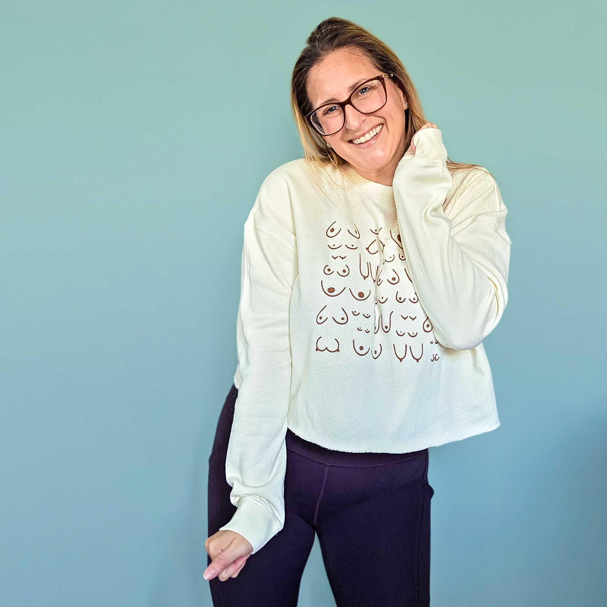 Woman wearing a cream self-love crop sweatshirt with design and dark pants against a light blue background