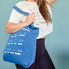 Person holding a blue tote bag with white text against a teal background
