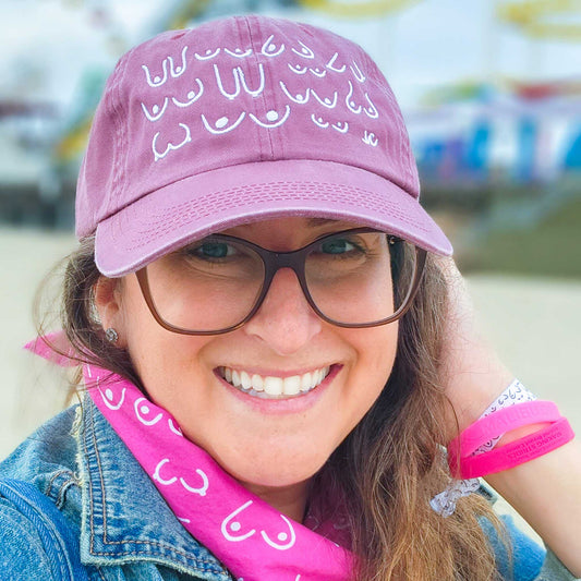 Woman wearing a pink cap and scarf with white text, smiling outdoors.